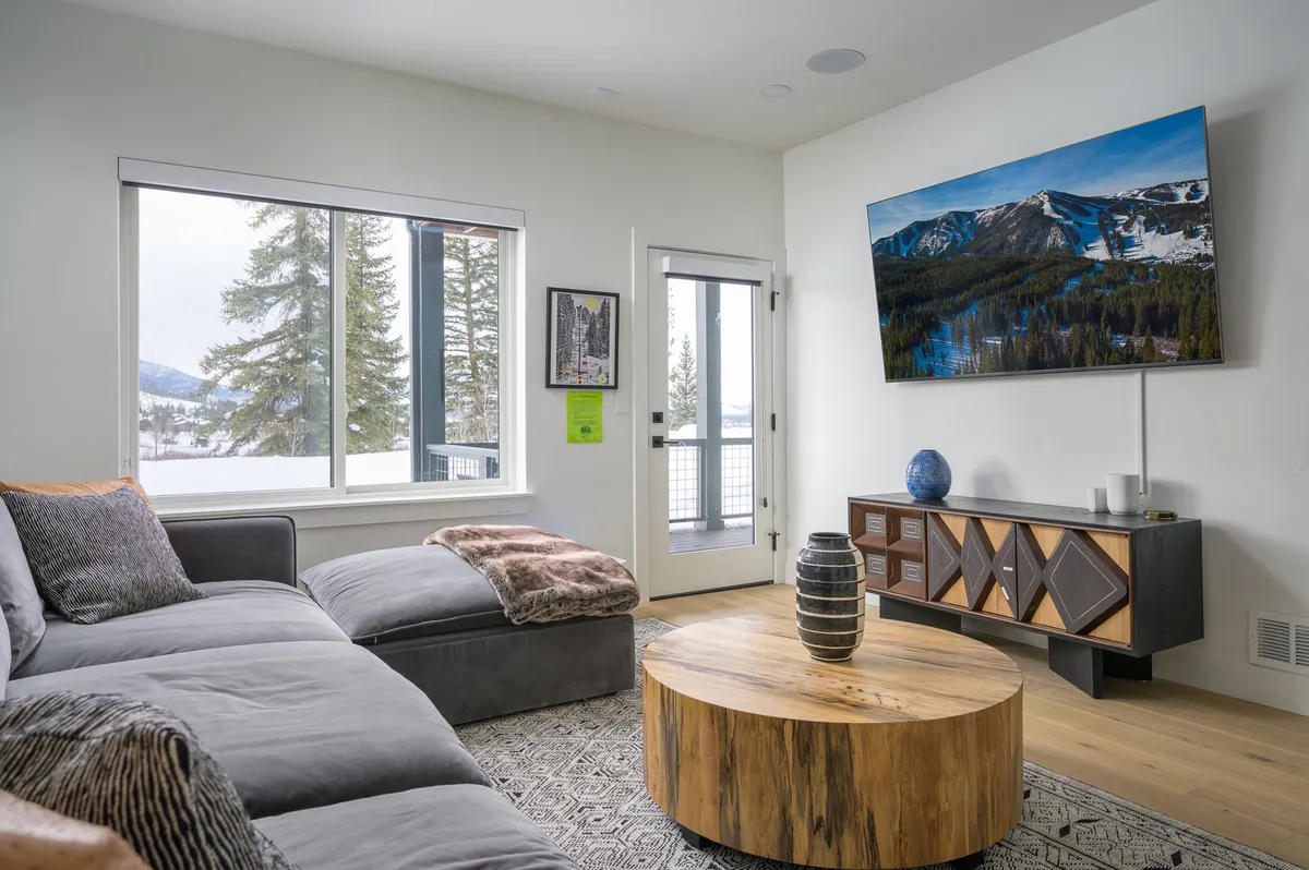 A spacious living area with a large sectional sofa, round wooden coffee table, and art on the wall. Large windows offer sweeping views of the mountains, and a flat-screen TV provides entertainment for group gatherings.
