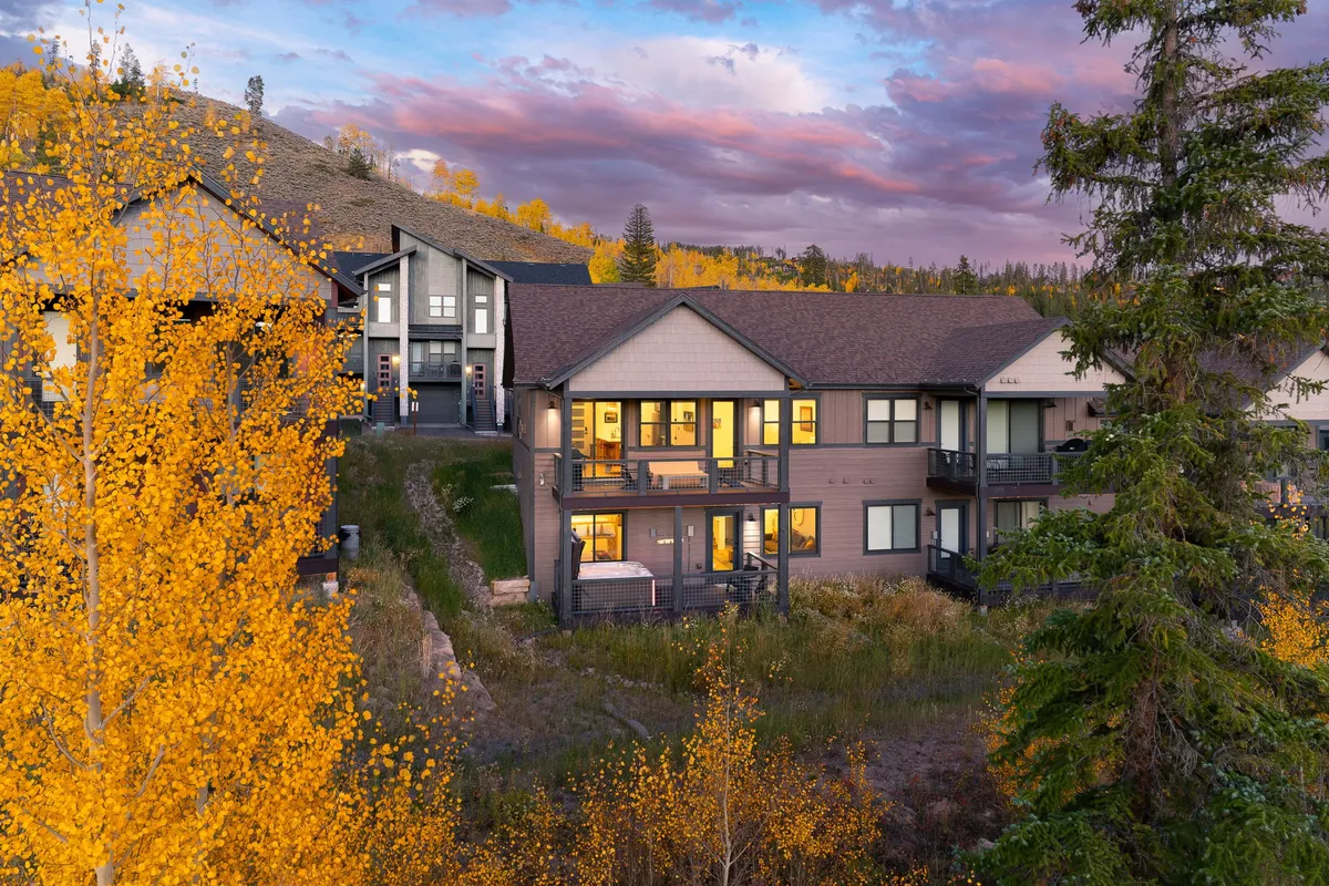 The image showcases the exterior of a luxurious chalet set against vibrant autumn foliage, with the beautiful mountain landscape of Fraser, Colorado in the background. The facade is inviting, featuring balconies perfect for relaxing or enjoying the panoramic views. The sky is dotted with soft clouds, creating a tranquil and welcoming mood.