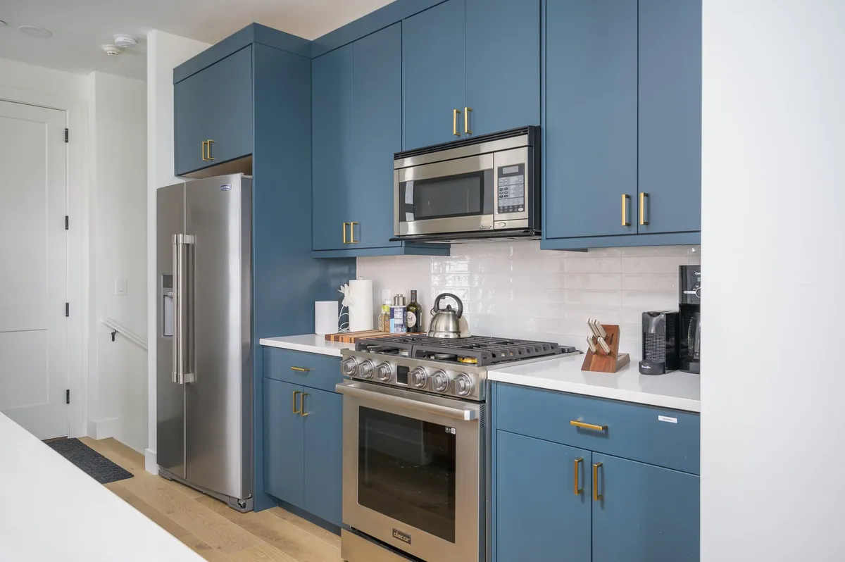 A close-up of the kitchen’s blue cabinetry, white countertops, stainless steel appliances, and countertop appliances such as a kettle and coffee maker. The space embodies a functional yet stylish atmosphere, suggesting both gourmet cooking and daily comforts like coffee-making.