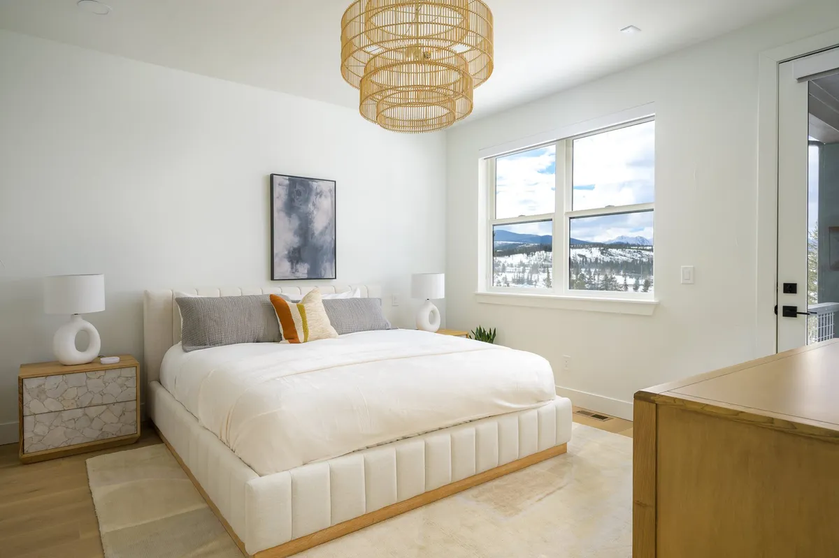 A sunlit bedroom featuring a plush bed adorned with decorative pillows, a modern hanging light fixture, and a large window offering stunning views of the Rocky Mountains. The room exudes tranquility, with soft textiles and a welcoming atmosphere, perfectly suited for restful sleep or peaceful reading.