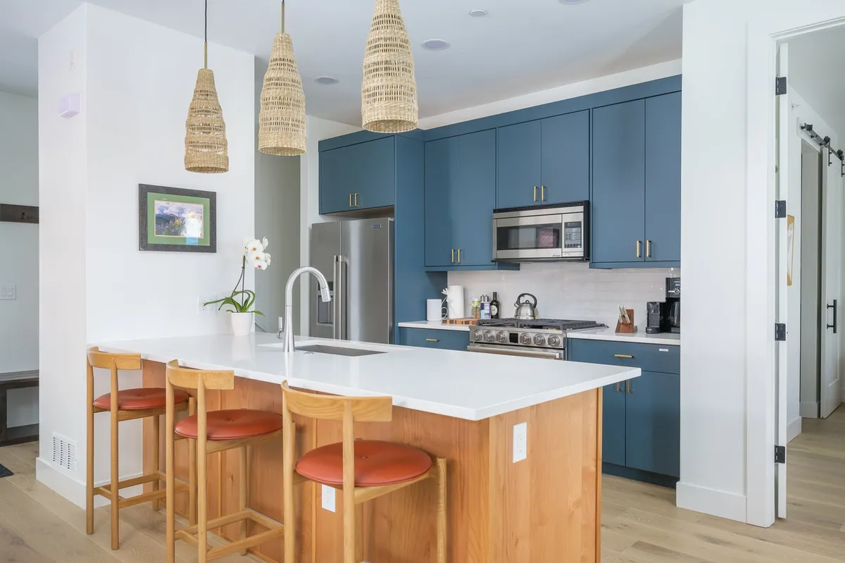 A modern kitchen featuring a spacious island countertop with bar stools, elegant pendant lights overhead, striking blue cabinetry, and high-end stainless steel appliances. The setup highlights ample prep space and a welcoming area for casual dining or guest interaction.