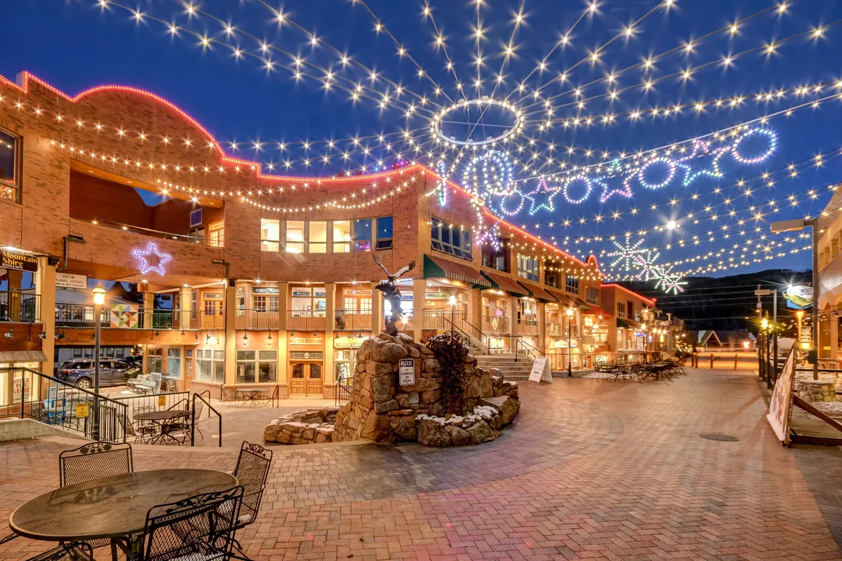 An enchanting nighttime scene featuring twinkling string lights, decorative stars, a stone fountain, and an outdoor seating area. The setting is festive and inviting, suggesting both the possibility of evening dining and the magical ambiance of downtown events or strolls.