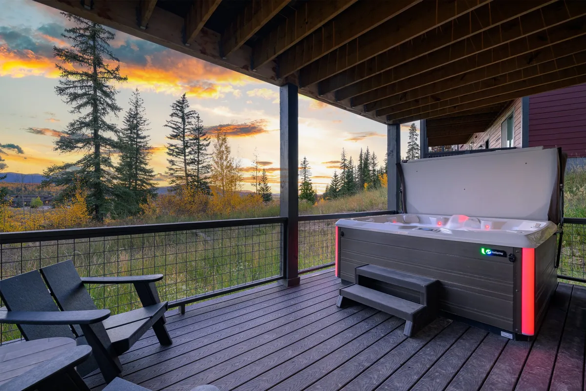 This image captures a serene exterior view of the covered patio with a hot tub, surrounded by natural forest and mountain scenery. Deck seating is visible, and the scene is set at sunset, highlighting a peaceful, relaxing atmosphere.