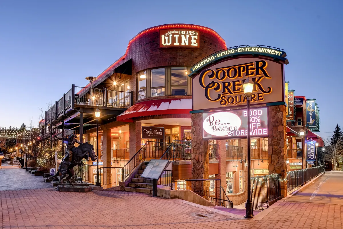 This vibrant exterior image captures the entrance to Cooper Creek Square, a lively shopping and dining hub in downtown Winter Park. Visible are shopfronts, a prominent Wine Shop, a sculpture of a horse, and a welcoming sign, with a stairway leading up to additional shops. The scene is bustling, evoking the dynamic energy of Winter Park's downtown scene.