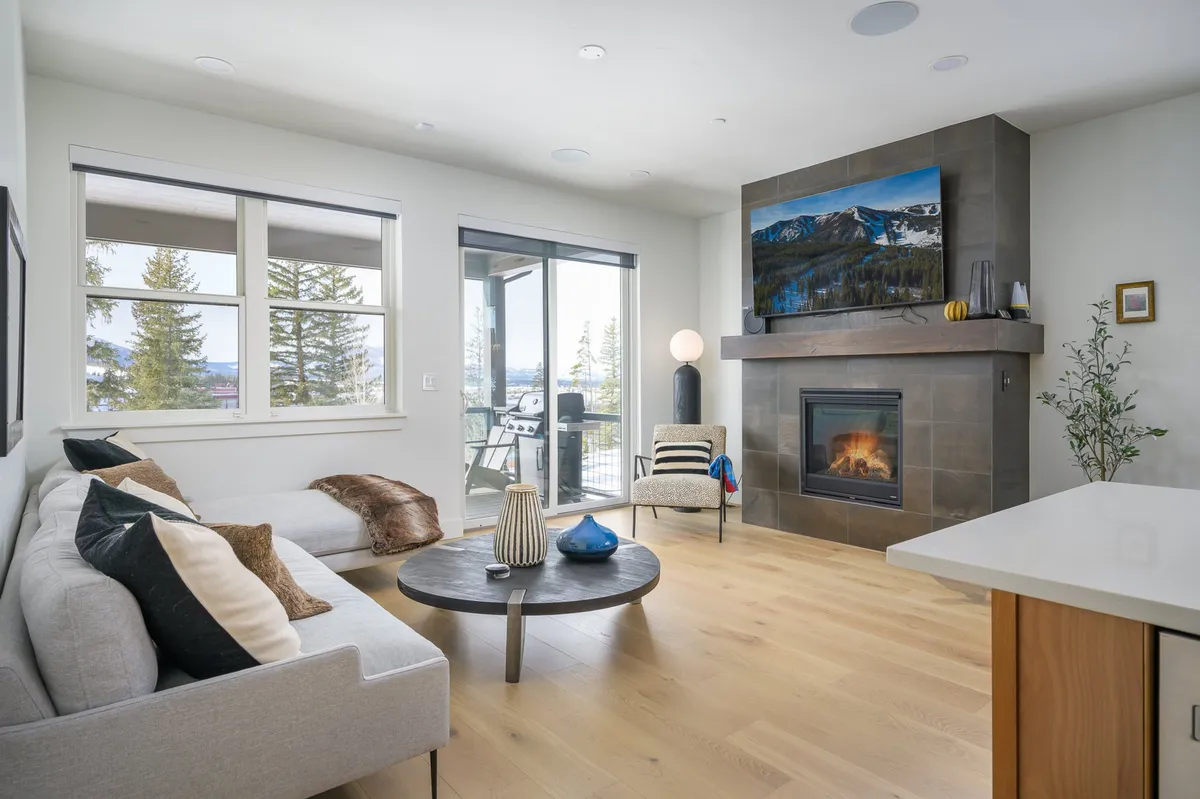 This image features a comfortable sofa set around a modern fireplace, with a wall-mounted TV and large windows that reveal a panoramic mountain view. The room is bright and airy, perfectly capturing the essence of a great room designed for gatherings and relaxation.